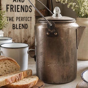 May include: A weathered, cylindrical metal coffee percolator with a wire bail handle, a side handle, and a clear glass knob on the lid. The surface shows signs of age and patina. The percolator is positioned near white enamel mugs and bread.