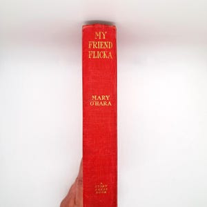 May include: A red hardcover book with the title "My Friend Flicka" by Mary O'Hara in gold lettering. The book is a Story Press Book. The spine is visible.