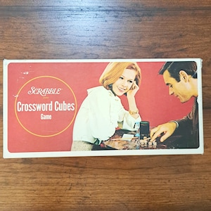 May include: A vintage Scrabble Crossword Cubes game box. The box features a red background with a woman and a man playing the game. The Scrabble logo and game title are in a yellow circle. The box is rectangular and has a white border.
