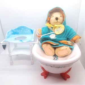 May include: A teddy bear wearing a teal hooded robe with orange trim sits in a white bathtub with red feet. A small white changing table with a blue top is to the left. The bear holds a spoon.
