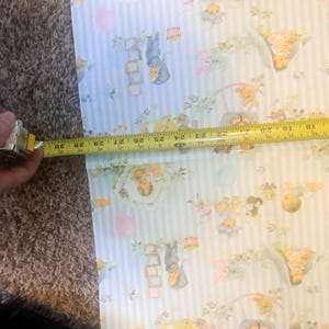May include: Light blue and white striped wrapping paper featuring a repeating pattern of illustrated babies, alphabet blocks, and balloons. A yellow measuring tape is placed across the paper, showing a width of approximately 12 inches.