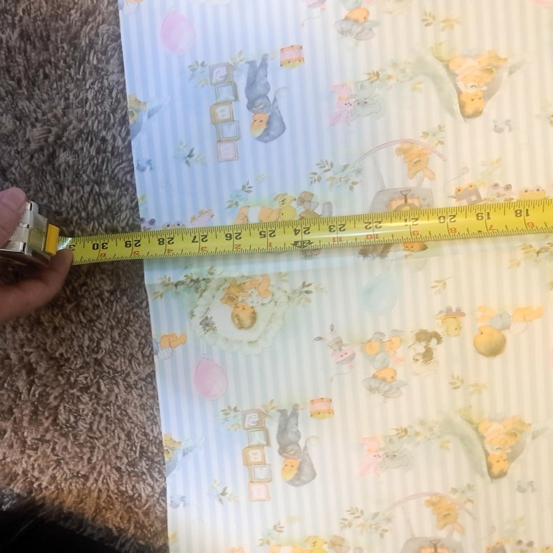 May include: Light blue and white striped wrapping paper featuring a repeating pattern of illustrated babies, alphabet blocks, and balloons. A yellow measuring tape is placed across the paper, showing a width of approximately 12 inches.