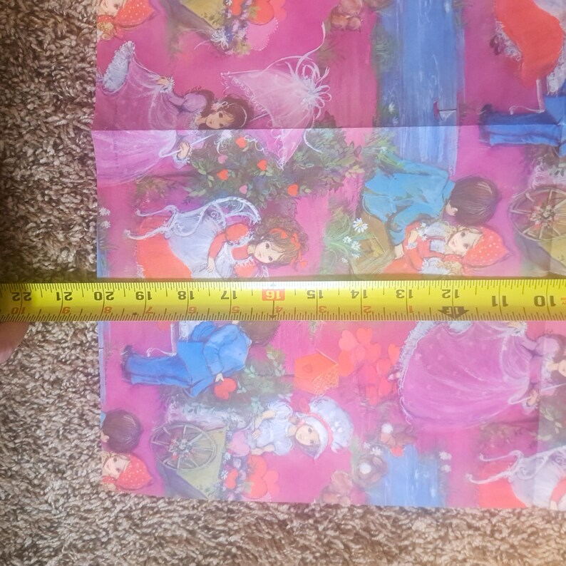 May include: A pink fabric featuring a repeating illustration of children in various outfits, including red hoods and blue suits, alongside small carts and floral elements. A yellow measuring tape is placed across the fabric, showing a width of 12 inches.