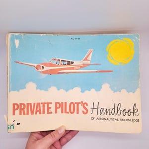 May include: Vintage "Private Pilot's Handbook" book. The cover features a red and white airplane illustration against a blue sky with a yellow sun and white clouds. The title is in red and black text.