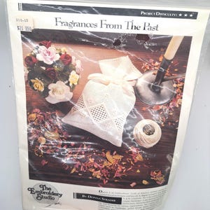 May include: A white embroidery project kit with a white sachet bag, a spool of white thread, and a bouquet of white and pink roses. The kit is titled "Fragrances From The Past" and is by Donna Strader. The project difficulty is rated as three stars.