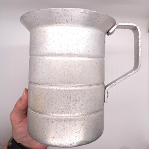 May include: A silver aluminum measuring cup with a handle. The cup has graduated markings and is designed for measuring liquids.