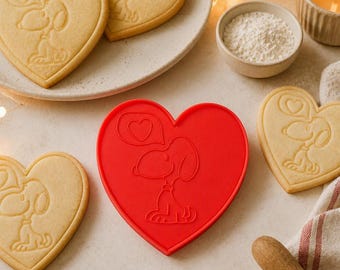 May include: A bright red heart-shaped cookie cutter featuring an embossed design of the cartoon character Snoopy sitting and a heart-shaped speech bubble above. The cutter is surrounded by several heart-shaped sugar cookies with the same design.