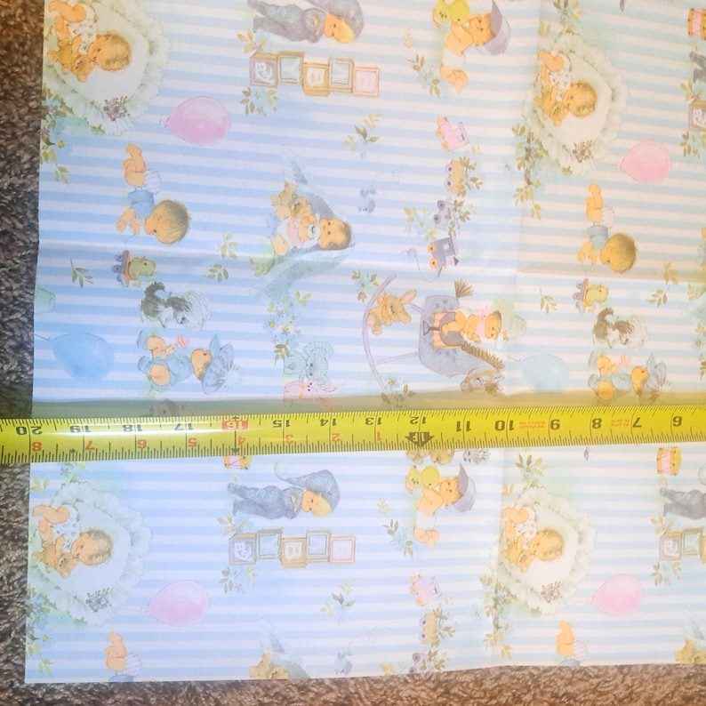 May include: Light blue and white striped fabric featuring a repeating pattern of babies, alphabet blocks spelling BABY, balloons, and small toys. A yellow measuring tape is placed across the fabric, showing a width of approximately 20 inches.
