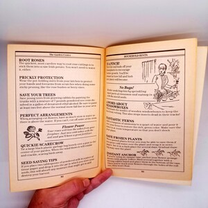 May include: Open book with gardening tips and household hints. The pages are yellowed, with text and illustrations. Topics include root roses, prickly protection, and saving trees. A hand is holding the book.