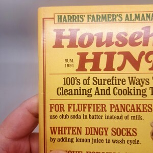 May include: A close-up of a vintage book cover titled "Harris' Farmer's Almanac Household Hints." The cover is yellow with brown text, including the year "1991." The book offers tips on cleaning and cooking, with examples like "For Fluffier Pancakes."
