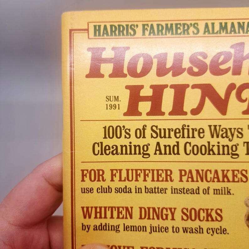 May include: A close-up of a vintage book cover titled "Harris' Farmer's Almanac Household Hints." The cover is yellow with brown text, including the year "1991." The book offers tips on cleaning and cooking, with examples like "For Fluffier Pancakes."