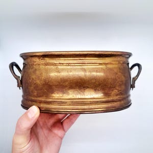 May include: A vintage, antique brass planter with a weathered, golden finish. The rectangular planter features two curved handles and horizontal ridges around the body. The planter is held by a hand against a white background.