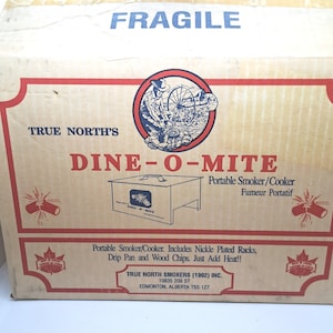 May include: A cardboard box with the text "FRAGILE" in blue. The box features the words "TRUE NORTH'S DINE-O-MITE" in red, with a graphic of a smoker. The box also states "Portable Smoker/Cooker" and "Includes Nickle Plated Racks, Drip Pan and Wood Chips."