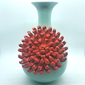 May include: A light mint green ceramic vase featuring a prominent, three-dimensional, glossy orange flower attached to the front. The vase has a smooth, rounded body and a slender, flared neck, creating a striking contrast with the textured petals.