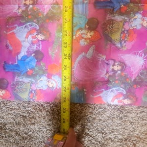 May include: A pink fabric featuring a repeating illustration of children in vintage clothing, including figures with umbrellas and carts. A yellow measuring tape is placed vertically over the fabric, showing a length of approximately 13 inches.
