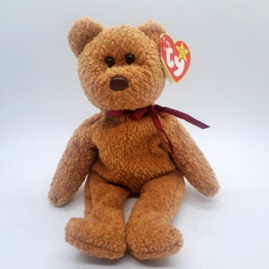 May include: A brown teddy bear with a red ribbon around its neck and a Ty heart-shaped tag. The plush toy has dark brown eyes and a brown nose. The bear is sitting on a white surface.