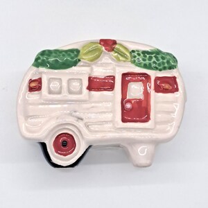 May include: A white ceramic camper with red trim and a green garland on top. The camper has a red door and three windows.