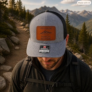 May include: A heather gray and black trucker hat featuring a rectangular brown leather patch with a mountain graphic and the text I RESCUE ROCKS TRAPPED IN MOUNTAINS. A black and white sticker on the brim displays the Richardson 112 brand logo.
