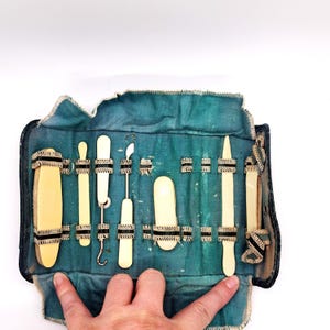 May include: An antique medical instrument kit. The kit includes various ivory-colored tools, such as probes and spatulas, secured within a teal fabric case. The case is trimmed with black and white ribbon and has a dark blue border.