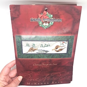May include: A Christmas-themed artwork titled "Dashing Through the Snow" featuring otters in a snowy landscape. The artwork is framed with a green border and includes the text "When Friends Gather" and "Pine Needles".