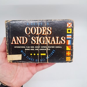 May include: A vintage book titled "Codes and Signals" with a black cover and distressed edges. The cover features the title in large orange letters, along with information about flag codes, Morse code, and sign language. Colorful flags are displayed on the right side.