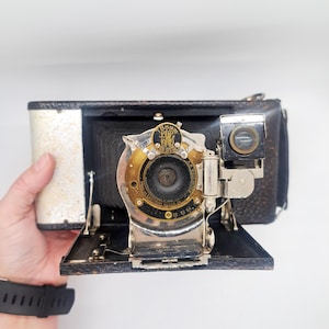 May include: Vintage black and silver Kodak camera with a leather exterior and a lens with gold and black accents. The camera has a viewfinder and a bellows design, showcasing its classic aesthetic.