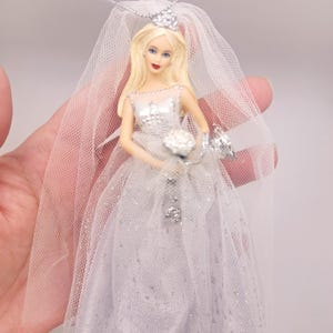 Peut inclure: Une poupée de mariée miniature vêtue d'une robe de mariée blanche avec un voile et un diadème. La poupée tient un bouquet et un petit sac à main. La robe a un revêtement scintillant. La poupée a les cheveux blonds et du rouge à lèvres rouge.