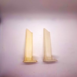May include: Two off-white plastic pieces with a rectangular shape and a slanted top. The pieces have a base and are standing upright against a white background. The plastic has a slightly translucent appearance.
