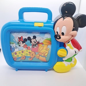 May include: A vintage blue Mickey Mouse toy TV with a handle and a yellow latch. The screen shows Mickey and Minnie Mouse on a train. Mickey Mouse is a 3D figure on the side, wearing a red jacket, yellow shoes, and white gloves.