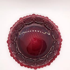 May include: A deep red glass plate with a scalloped edge and ornate embossed detailing. The plate has a glossy finish and a slightly raised center. The design features repeating oval and dot patterns around the rim.