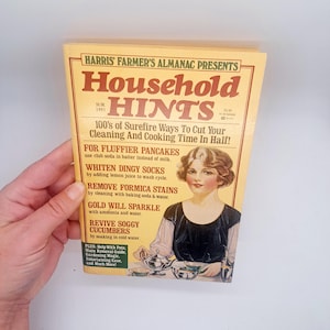 May include: A vintage "Household Hints" almanac from 1991, held by a hand. The cover features a woman in a black dress and the text "100's of Surefire Ways To Cut Your Cleaning And Cooking Time In Half!" in red and brown.