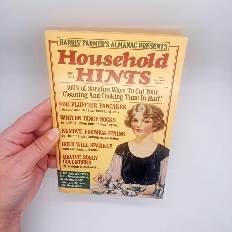 May include: A vintage "Household Hints" almanac from 1991, held by a hand. The cover features a woman in a black dress and the text "100's of Surefire Ways To Cut Your Cleaning And Cooking Time In Half!" in red and brown.