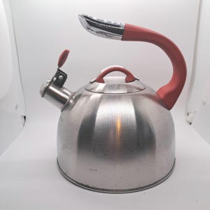 May include: A stainless steel stovetop kettle with a brushed metal finish. It features a curved handle with a coral-colored silicone grip, a matching coral-colored lid knob, and a whistle spout with a coral-colored lever for opening the spout.