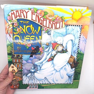 May include: A children's book cover featuring a colorful illustration of the Snow Queen holding a young child in her arms. The title of the book is "The Snow Queen" by Hans Christian Andersen. The cover is illustrated by Mary Engelbreit.