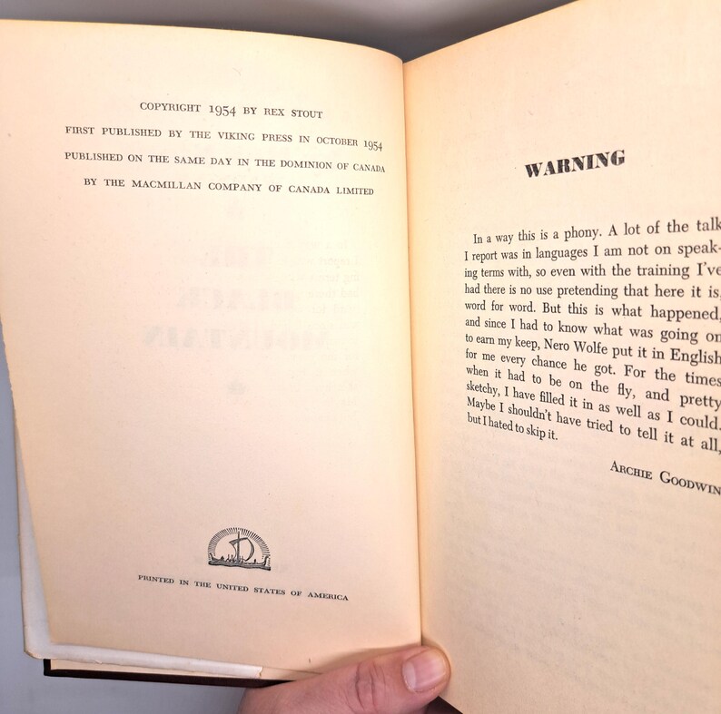 Puede incluir: Libro abierto con texto, incluyendo "COPYRIGHT 1954 BY REX STOUT" y "WARNING". Las p&aacute;ginas son de color blanquecino con texto negro. El libro est&aacute; abierto en una p&aacute;gina con texto y un peque&ntilde;o emblema en la parte inferior. Una mano sostiene el libro.
