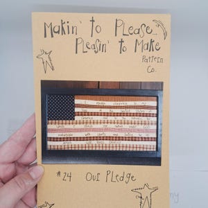 May include: A framed textile art piece featuring a patchwork American flag design. The flag includes the text of the Pledge of Allegiance. The artwork is displayed on a tan background with handwritten text and star illustrations.