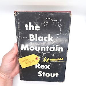Puede incluir: Un libro de tapa dura vintage titulado "The Black Mountain" de Rex Stout. El libro tiene una cubierta negra con letras blancas y una etiqueta amarilla que dice "Nero Wolfe novel". El libro est&aacute; sostenido por una mano.