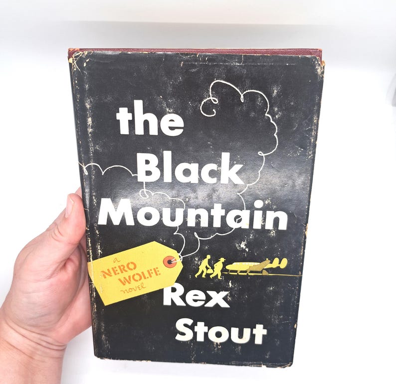 Puede incluir: Un libro de tapa dura vintage titulado "The Black Mountain" de Rex Stout. El libro tiene una cubierta negra con letras blancas y una etiqueta amarilla que dice "Nero Wolfe novel". El libro est&aacute; sostenido por una mano.