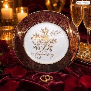 May include: A round ceramic plate with a white center and a wide, dark burgundy rim featuring a gold floral pattern. The center displays a gold illustration of a rose with the text 40th Anniversary in a cursive script font.