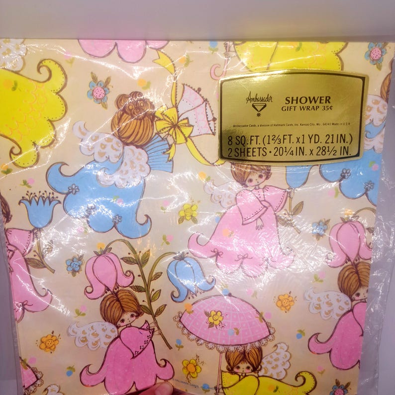 May include: A package of Ambassador shower gift wrap featuring a cream background with a repeating pattern of angels in pink and blue dresses, yellow umbrellas, and flowers. The gold label reads: Ambassador SHOWER GIFT WRAP 8 SQ. FT. (1 2/3 FT. x 1 YD. 21 IN.) 2 SHEETS 20 1/4 IN. x 28 1/2 IN.