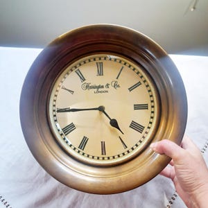 May include: A round, antique-style clock with a bronze-colored frame. The clock face is cream-colored with black Roman numerals and hands. The text "Harrington & Co. LONDON" is printed on the clock face.