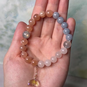 May include: A multi-colored beaded bracelet with a crescent moon charm. The bracelet is made with light pink, white, and blue beads. The charm is made of clear glass with iridescent colors.
