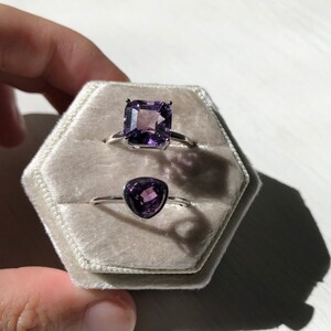 May include: Two rings with purple gemstones in a white velvet ring box. The larger ring has a square-shaped gemstone, while the smaller ring has a triangular gemstone.