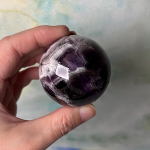 May include: A polished amethyst sphere, displaying a mix of deep purple and white bands. The gemstone ball is held in a hand, with light reflecting off its smooth surface. This decorative sphere is a unique home decor item.