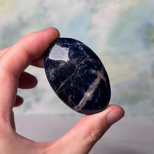 Puede incluir: Una piedra de palma de sodalita azul lisa de forma ovalada con vetas blancas y doradas.