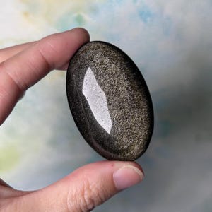 May include: Oval-shaped, polished golden obsidian stone. The stone has a dark, reflective surface with shimmering gold inclusions. A large, bright white, irregular shape reflects light on the surface. The stone is held between fingers.