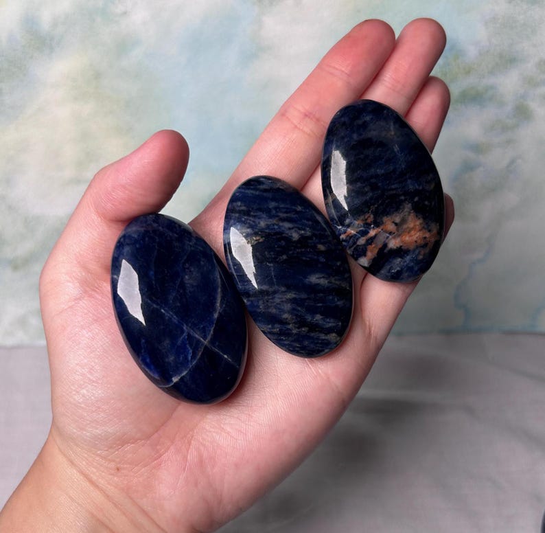 Puede incluir: Tres piedras de sodalita azul lisas de forma ovalada con vetas blancas y marrones. Las piedras est&aacute;n pulidas y tienen un acabado brillante.