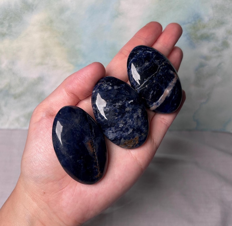 Puede incluir: Tres piedras lisas de sodalita azul de forma ovalada y pulidas. Las piedras son de color azul oscuro con vetas blancas.