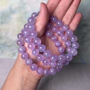 May include: A strand of lavender amethyst beads, with a slightly iridescent finish, is wrapped around a hand. The beads are round and smooth.