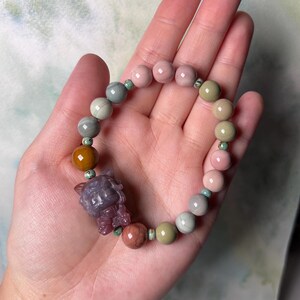 May include: A multi-coloured gemstone bracelet with a purple jade charm. The bracelet features beads in shades of pink, green, brown, and white.